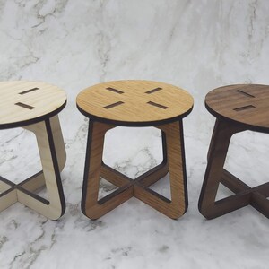 May include: Three small wooden stools in varying shades of light, medium, and dark brown. Each stool has a round seat with four cut-out slots and an X-shaped base. The stools are displayed on a marble-patterned surface.
