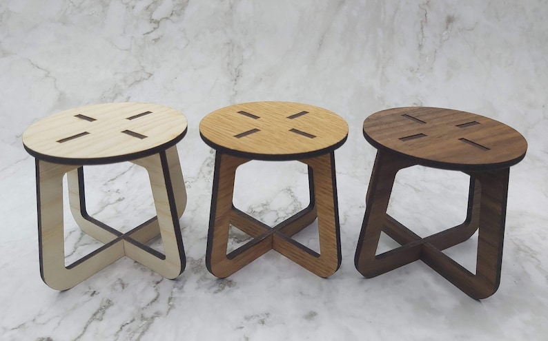 May include: Three small wooden stools in varying shades of light, medium, and dark brown. Each stool has a round seat with four cut-out slots and an X-shaped base. The stools are displayed on a marble-patterned surface.