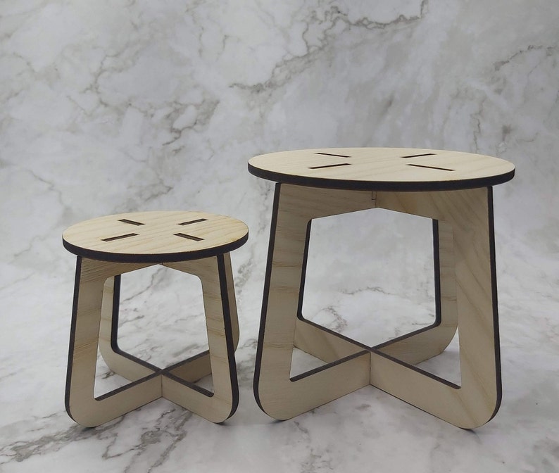 May include: Two wooden stools of different sizes. The stools have a natural wood finish with dark brown edges. Each stool has a round top with four evenly spaced slots and a geometric base with curved legs.