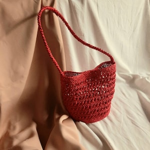 May include: A red crocheted shoulder bag with a braided handle. The bag has an open weave design and a rounded shape. The bag is made of a red yarn and is displayed against a backdrop of draped fabric.