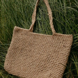 May include: A large, brown, woven tote bag made of natural fibers. The bag has two long straps and is sitting on a bed of green grass.