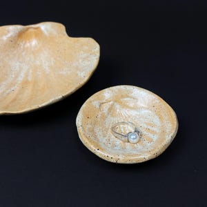 May include: Two ceramic seashell-shaped dishes in shades of tan and cream. A small dish holds a silver ring with a pearl. The dishes have a speckled texture and are set against a black background.