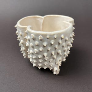 Könnte beinhalten: Ein weißer Keramik-Aschenbecher mit einem einzigartigen, strukturierten Design. Der Aschenbecher hat eine dreilappige Form und ist mit kleinen, spitzen Stacheln bedeckt. Das Innere ist glatt, im Gegensatz zur rauen Textur des Äußeren. Auf dunklem Hintergrund.