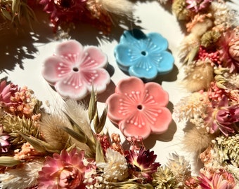 May include: Three ceramic flower-shaped incense holders in pink and blue. The holders are arranged on a white surface surrounded by a wreath of dried flowers.