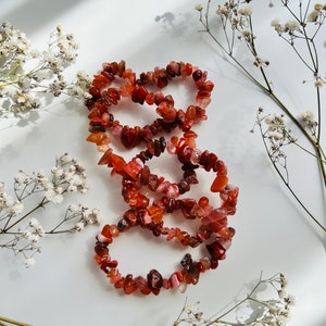 Op de afbeelding: Drie strengen carneol-chip kralen armbanden in tinten van rood en oranje, gerangschikt in een stapel op een witte achtergrond met witte bloemen.
