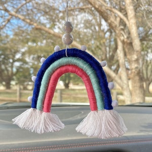 May include: A rainbow-colored yarn car air freshener with white pom-poms and a white tassel on each end. The air freshener is hanging from the rearview mirror of a car.