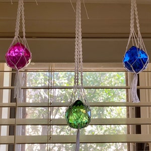 May include: Three crystal ball ornaments hanging from macrame plant hangers. The balls are pink, blue, and green.