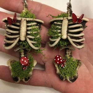 May include: A pair of skeleton earrings with moss and red mushrooms. Each earring features a small red butterfly.