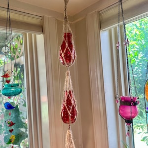 May include: Red glass hanging planters suspended from the ceiling. The planters are encased in a woven net and have a tassel at the bottom. Other hanging glass decorations are visible, including a teal bird feeder and a pink candle holder.