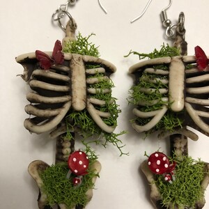 May include: A pair of skeleton earrings with moss, mushrooms, and butterflies. The earrings are made of a white and brown plastic material and have silver hooks.