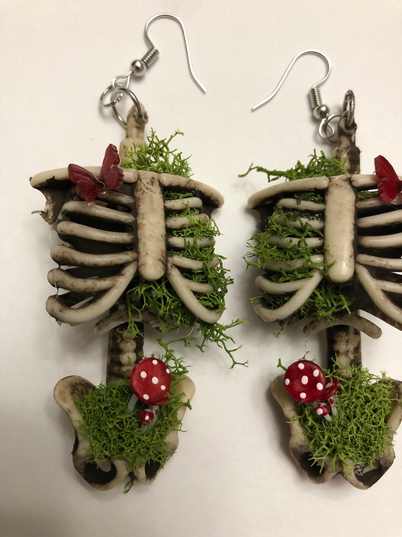 May include: A pair of skeleton earrings with moss, mushrooms, and butterflies. The earrings are made of a white and brown plastic material and have silver hooks.