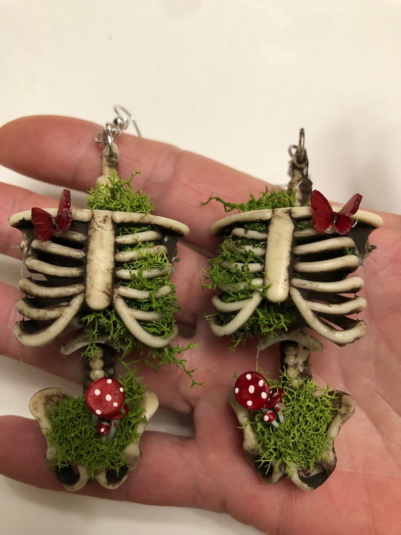 May include: A pair of skeleton earrings with moss and red mushrooms. Each earring features a small red butterfly.