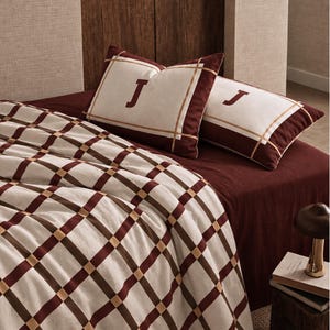 May include: A bedding set featuring a cream-coloured blanket with a dark red and tan grid pattern. Two matching rectangular pillows are included, each displaying a dark red letter J centred on a cream panel framed by dark red and tan borders.