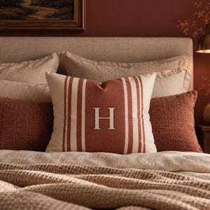 May include: A square throw pillow featuring a central rust-colored panel with vertical cream stripes and a large, centered cream-colored letter H. The pillow is made of a textured fabric and is positioned among other neutral and rust-colored bedding.