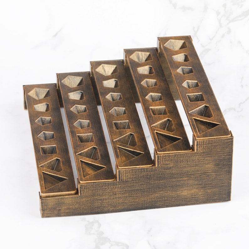 Peut inclure: Un organisateur en bois de couleur bronze, en forme d'escalier, avec cinq sections en gradins. Chaque section comporte des d&eacute;coupes g&eacute;om&eacute;triques et est con&ccedil;ue pour contenir de petits objets. L'organisateur repose sur une surface en marbre blanc.