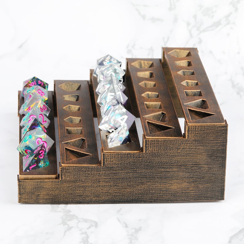 Peut inclure: Un pr&eacute;sentoir &agrave; d&eacute;s en bois &agrave; plusieurs niveaux, teint&eacute; en marron fonc&eacute;, avec des d&eacute;coupes g&eacute;om&eacute;triques. Plusieurs jeux de d&eacute;s transparents color&eacute;s sont expos&eacute;s, pr&eacute;sentant diverses couleurs et motifs. Le pr&eacute;sentoir est con&ccedil;u pour ranger les d&eacute;s.