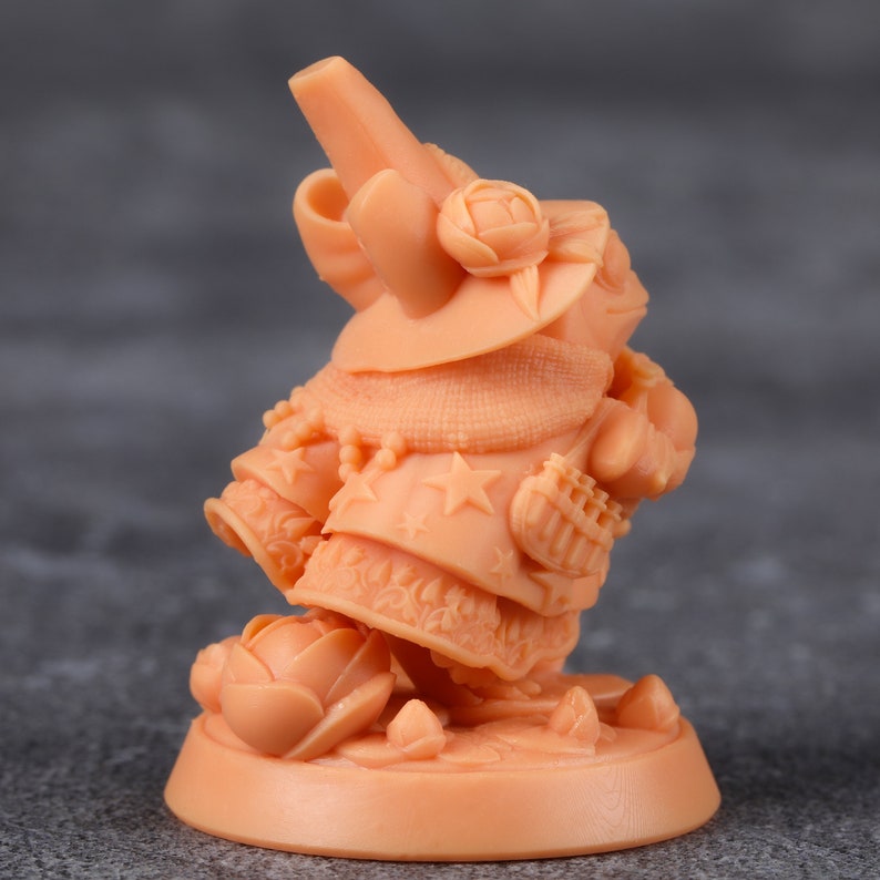 Op de afbeelding: Perzik kleurige beeldje van een fantasiefiguur, mogelijk een gnome of tovenaar, met een hoed, cape en bloemmotieven. De figuur staat op een ronde basis met een bloem aan zijn voeten. Details omvatten sterren en ingewikkelde patronen.