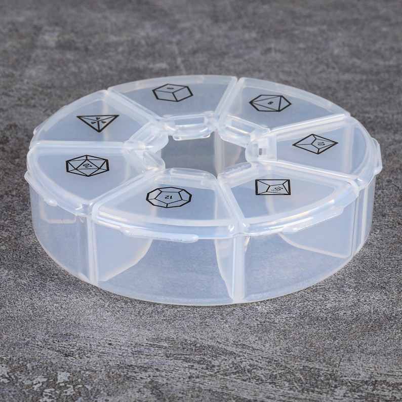 Puede incluir: Una caja de pastillas de pl&aacute;stico transparente con siete compartimentos. Cada compartimento est&aacute; etiquetado con un n&uacute;mero diferente, que representa los lados de un dado.