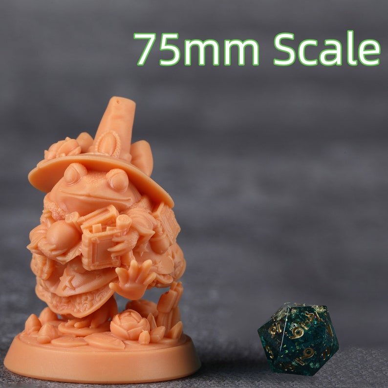 Op de afbeelding: Perzik kleurige beeldje van een kikker heks, met een hoed en een boek. De tekst "75mm Scale" is zichtbaar. Een veelzijdige dobbelsteen met gouden cijfers staat naast de kikker.