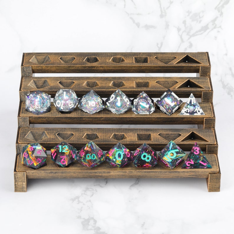 Peut inclure: Un pr&eacute;sentoir en bois pour d&eacute;s contient une collection de d&eacute;s poly&eacute;driques iris&eacute;s. Les d&eacute;s sont transparents avec des paillettes color&eacute;es et des faces num&eacute;rot&eacute;es. Le pr&eacute;sentoir est en bois brun fonc&eacute;.