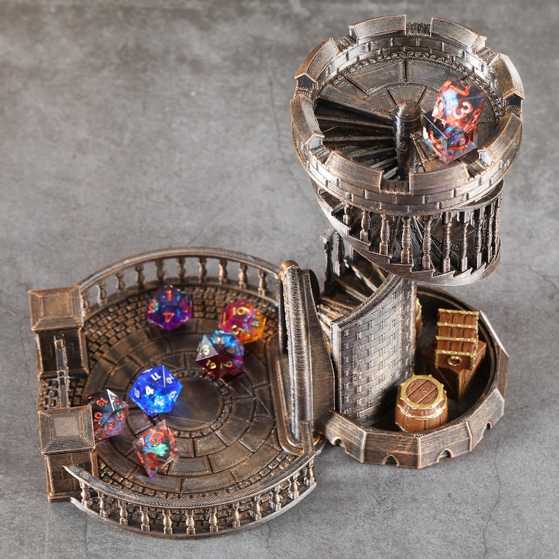 May include: A bronze-colored tabletop game tower with a spiral staircase and a circular platform. The tower has a removable top section that can be used to roll dice. The platform has a circular design and is surrounded by a railing. There are six multi-colored dice on the platform.