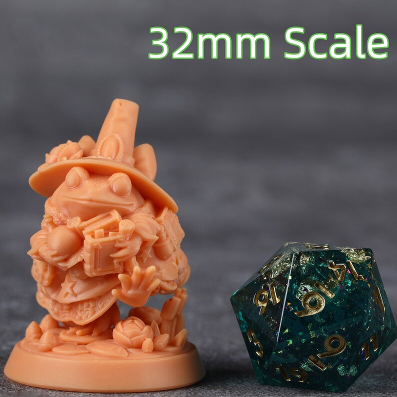Op de afbeelding: Een perzik-kleurige kikkerfiguur van 32 mm, met een hoed en een boek, naast een groen en goud twintigzijdig dobbelsteen. Het beeldje is gedetailleerd met bloemaccenten en een grillig ontwerp, voor tabletop games.