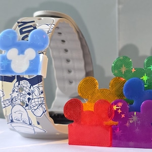 May include: A selection of colourful Mickey Mouse-shaped accessories, including red, orange, yellow, green, blue, purple, and white. Two Disney-themed watch straps are also visible, one featuring Stitch and the other with cartoon characters.