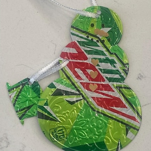 May include: A handmade snowman ornament crafted from recycled aluminum cans. The snowman features a green and white body with red accents and small heart details. A silver ribbon is attached for hanging. The ornament has a unique, textured appearance.
