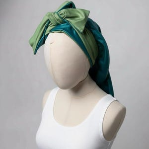 May include: A two-toned head wrap in shades of green and teal, tied in a bow on top. The head wrap is made of a silky material and is displayed on a mannequin head. The mannequin is wearing a white tank top.