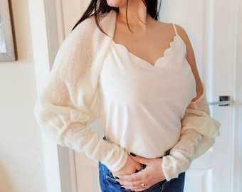 May include: A cream-colored, open-front shrug with long sleeves, paired with a white camisole and blue jeans. The shrug has a delicate, open knit texture. The camisole features a scalloped neckline. The woman is smiling.