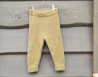 May include: Olive green baby pants hanging on a gold hanger against a gray wooden wall. The pants are made of a soft, textured knit fabric. The hanger is suspended from a decorative metal hook.