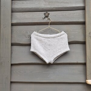 Puede incluir: Una prenda inferior de punto gris claro cuelga de una percha dorada. La prenda est&aacute; hecha de un material suave y texturizado. La percha est&aacute; suspendida de un gancho decorativo en una pared de madera gris. La prenda parece ser un pantal&oacute;n corto.