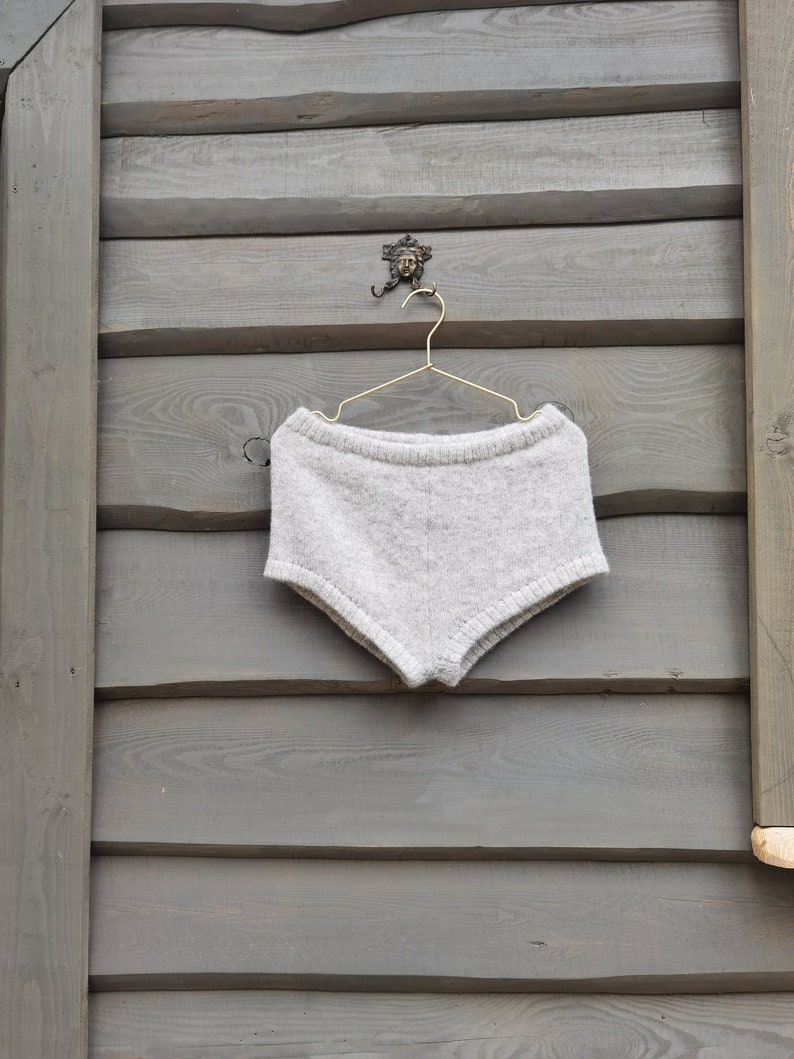 Puede incluir: Una prenda inferior de punto gris claro cuelga de una percha dorada. La prenda est&aacute; hecha de un material suave y texturizado. La percha est&aacute; suspendida de un gancho decorativo en una pared de madera gris. La prenda parece ser un pantal&oacute;n corto.