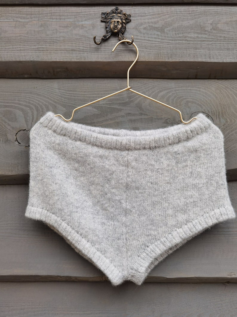 Puede incluir: Un short tejido de color gris claro, exhibido en una percha dorada. El short est&aacute; hecho de un material suave y texturizado. El fondo es una pared de madera gris desgastada con un gancho decorativo de metal.
