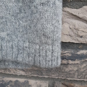 Puede incluir: Primer plano de una tela de punto gris, que muestra la textura y el tejido del material. La tela, probablemente de un su&eacute;ter o prenda similar, tiene un pu&ntilde;o acanalado en el borde inferior. El color es gris jaspeado.