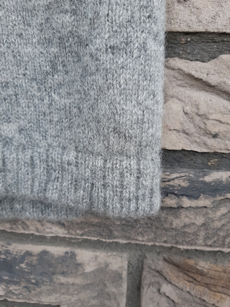 Puede incluir: Primer plano de una tela de punto gris, que muestra la textura y el tejido del material. La tela, probablemente de un su&eacute;ter o prenda similar, tiene un pu&ntilde;o acanalado en el borde inferior. El color es gris jaspeado.