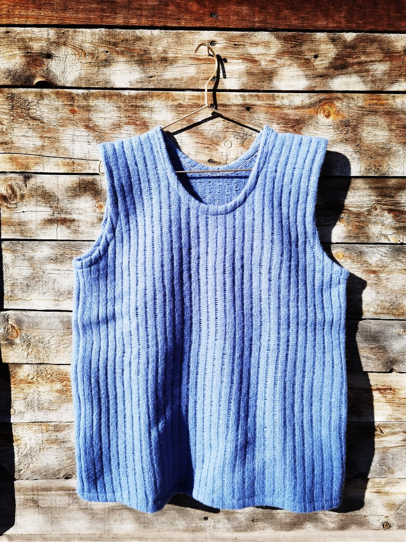 Puede incluir: Un chaleco azul sin mangas de punto colgado de una percha de madera. El chaleco tiene una textura acanalada y est&aacute; hecho de un material suave y c&oacute;modo.