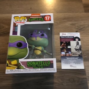 May include: A Donatello Pop! Vinyl Figure from the Teenage Mutant Ninja Turtles franchise. The figure is in its original packaging, with the number 17 on the box. A JSA authentication card is next to the figure.