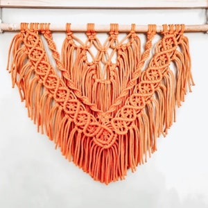 May include: A macrame wall hanging with a geometric design in a burnt orange colour. The hanging has a fringe border and is made with cotton cord.