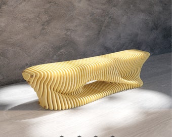Könnte beinhalten: Eine gelbe Holzbank mit einem einzigartigen, organischen, wellenförmigen Design. Die Bank besteht aus dünnen, geschichteten Holzstücken und hat einen Ausschnitt in der Mitte. Die Bank steht auf einem hellen Holzboden vor einer grauen Betonwand.