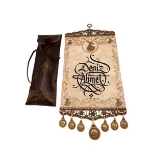 May include: A cream-coloured scroll with black calligraphy that reads "Deniz & Ahmet" in a decorative font. The scroll is framed with a gold metal border and has gold tassels hanging from the bottom. The scroll is rolled up and comes with a brown leather carrying case.