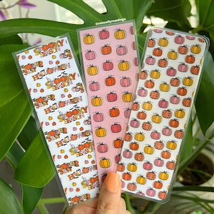 May include: Three Halloween themed bookmarks with a pumpkin pattern. The bookmarks are made of white cardstock with a clear plastic coating. The text on the bookmarks reads "It's Spooky Season" and "31st Halloween".
