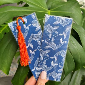 May include: Two blue and white koi fish pattern bookmarks with an orange tassel. The bookmarks are in clear plastic sleeves. The background is a large green leaf.