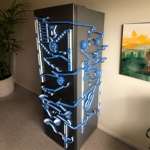 May include: A stainless steel refrigerator with a blue and white marble run attached to the front. The marble run is made of plastic and has a variety of curves, loops, and ramps.
