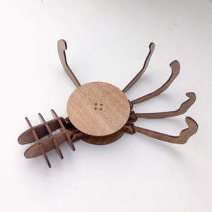 Könnte beinhalten: Ein Holzpuzzle in Form einer Krabbe mit rundem Körper und mehreren gebogenen Beinen. Das Puzzle besteht aus hellbraunem Holz und hat ein detailliertes, ineinandergreifendes Design. Der Körper der Krabbe hat vier kleine quadratische Aussparungen.