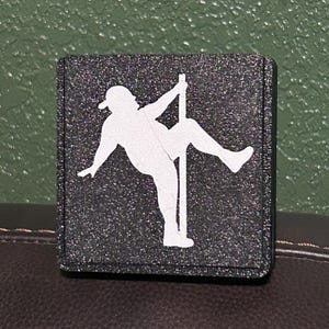 May include: A black square patch with a white silhouette of a person holding onto a pole. The patch has a glittery texture.