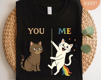 Op de afbeelding: Zwart T-shirt met een afbeelding van twee katten. Eén kat is bruin en chagrijnig, de andere is wit en vrolijk, met een regenboogstaart. De woorden "YOU" en "ME" staan boven de katten.