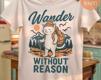 May include: Cream-colored t-shirt featuring a graphic of a white cat with a backpack, hiking stick, and hat, set against a mountain and forest backdrop. The text "Wander Without Reason" is displayed above and below the cat illustration.