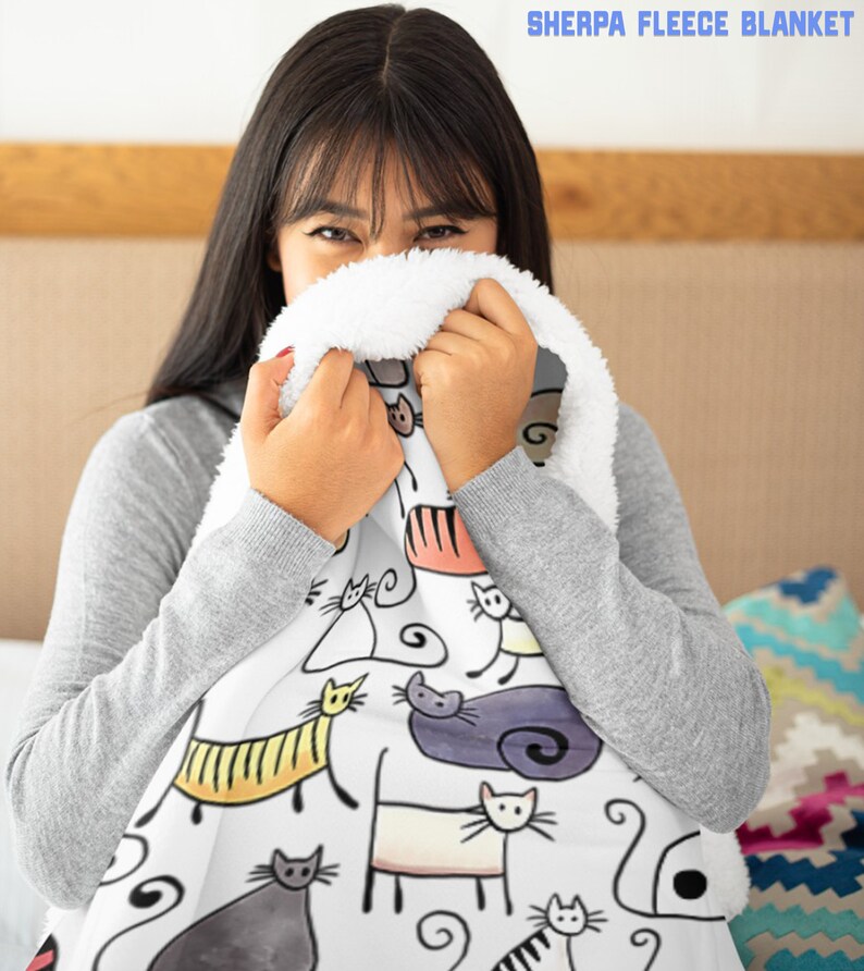 May include: A white sherpa fleece blanket with a colorful cat print. The blanket features a variety of cats in different poses and colors, including black, white, orange, and gray. The blanket is soft and cozy, perfect for snuggling up on the couch.