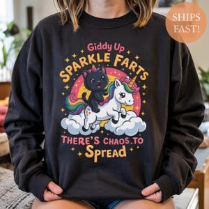 May include: Black sweatshirt with a colorful graphic of a black cat riding a unicorn on a cloud, with the text "Giddy Up Sparkle Farts There's Chaos To Spread". The design includes a rainbow and stars.